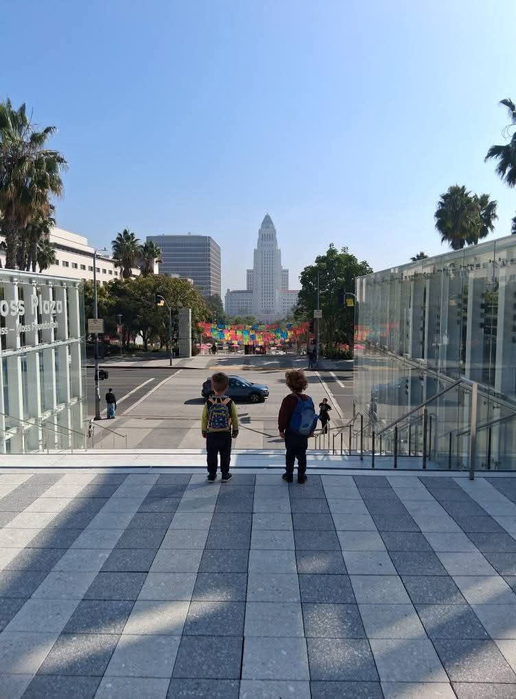 Two small children with backpacks walking toward a plaza with Los Angeles City Hall in the distance